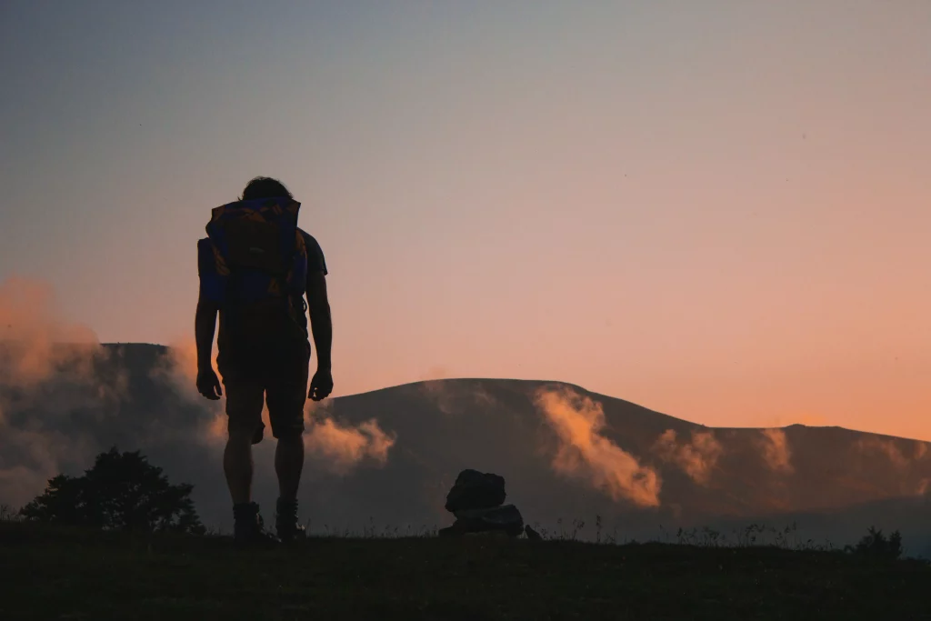 A photo with mountains. low clouds at sunrise, a silhoete of a man with a backpack heading out on a journey. 